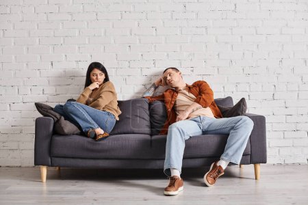 Photo for Young self-confident man talking to upset asian wife on couch in living room, divorce concept - Royalty Free Image