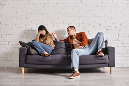 young emotional man talking to asian woman crying on couch in living room, divorce concept