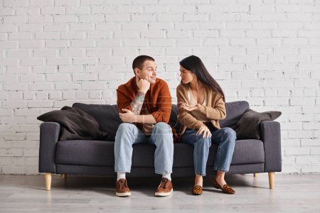 Familienscheidung, beleidigte Asiatin spricht mit lächelndem skeptischem Ehemann auf Couch im Wohnzimmer