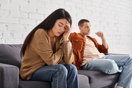 young depressed asian woman sitting with closed eyes near offended husband on couch in living room