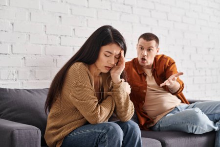 Wütender Mann streitet sich mit beleidigter asiatischer Frau, die mit geschlossenen Augen auf Couch zu Hause sitzt, Scheidung