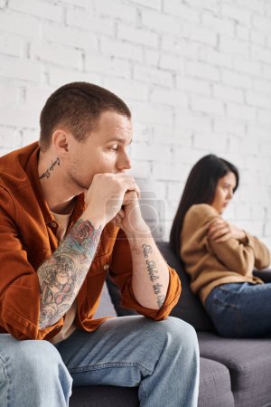 Téléchargez les photos : Frustré tatoué homme regardant offensé asiatique femme assis sur canapé à la maison, concept de divorce - en image libre de droit