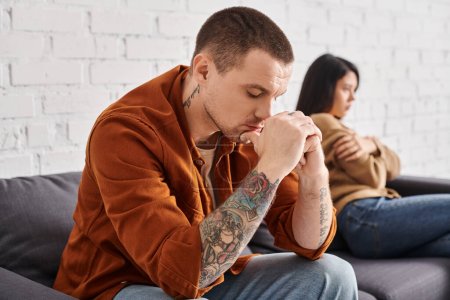 depressed tattooed man with closed eyes near offended asian wife on couch at home, divorce concept