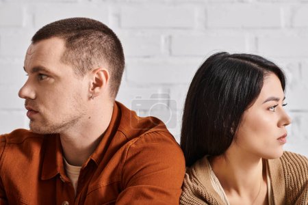 offended interracial couple sitting back to back and looking away in living room, divorce concept