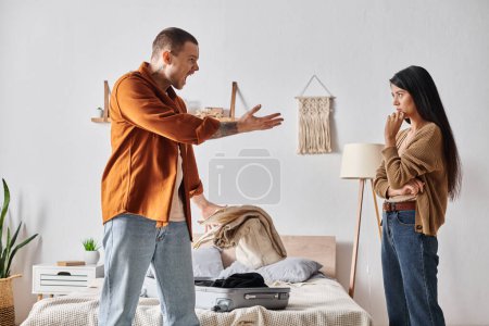 Téléchargez les photos : Jeune et en colère homme querelle à asiatique femme près de valise et biens dans chambre à coucher à la maison - en image libre de droit
