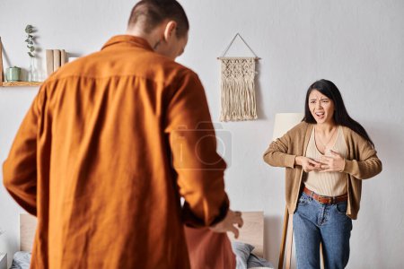 gereizte Asiatin streitet sich mit Mann, der mit Kleidung im Schlafzimmer steht, Familienstreit