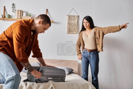 frustrated asian woman pointing away near husband packing suitcase at home, relationship trouble