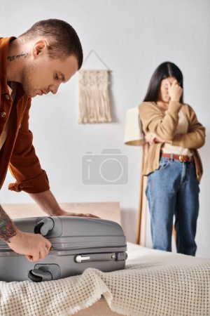 junger Mann packt Koffer in der Nähe einer depressiven asiatischen Frau, die zu Hause im Schlafzimmer weint, Scheidung der Familie