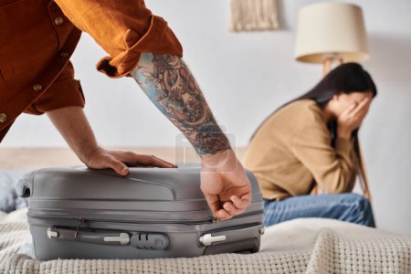 Photo for Young tattooed man packing suitcase near asian wife crying on bed at home, family misunderstanding - Royalty Free Image