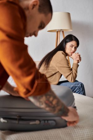 Téléchargez les photos : Stressé asiatique femme assis sur lit et regarder mari emballage valise, divorce concept - en image libre de droit
