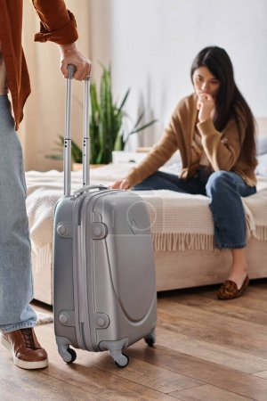 frustrierte asiatische Frau sitzt auf dem Bett, während ihr Mann mit Koffer das Haus verlässt, Scheidung