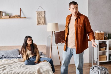Foto de Hombre con maleta y portátil saliendo de casa cerca molesto asiático esposa en dormitorio, divorcio concepto - Imagen libre de derechos