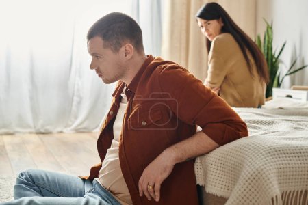 Photo for Young  frustrated man sitting on floor near upset asian wife, relationship difficulties concept - Royalty Free Image