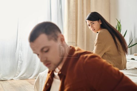 unzufriedene Stirnrunzeln asiatische Frau sieht jungen verärgerten Ehemann im Schlafzimmer zu Hause, Scheidungskonzept