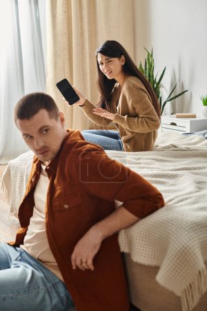 irritated asian woman with mobile phone looking at young  husband in bedroom at home, family trouble