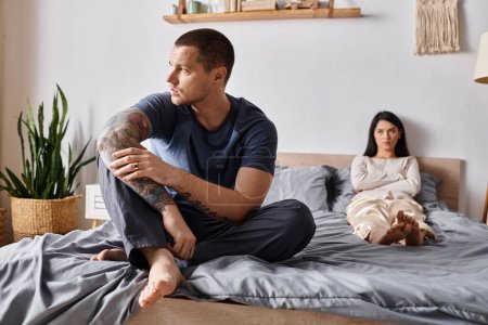 frustrated tattooed man sitting on bed near upset asian wife, family relationship difficulties