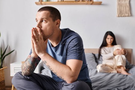 Téléchargez les photos : Jeune homme tatoué regardant loin tandis que assis sur le lit près offensé asiatique femme, concept de divorce - en image libre de droit