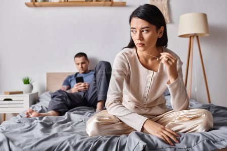 Téléchargez les photos : Bouleversé asiatique femme assis près de mari en utilisant smartphone dans chambre à coucher à la maison, problème familial - en image libre de droit