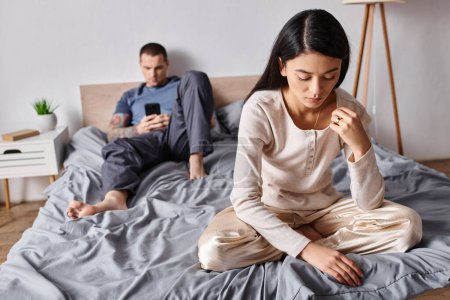 offended asian woman sitting near husband using smartphone in bedroom at home, family conflict