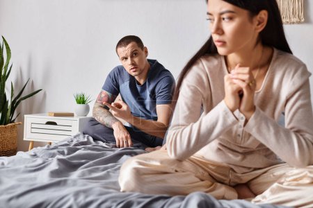 junger tätowierter Mann spricht mit verärgerter asiatischer Frau, während er zu Hause auf dem Bett sitzt, Scheidungskonzept