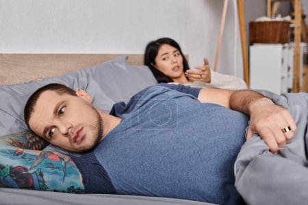 Téléchargez les photos : Jeune homme tatoué frustré regardant loin tout en étant couché sur le lit près de la femme asiatique, conflit familial - en image libre de droit