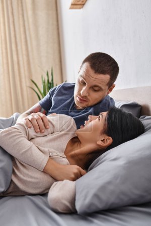 Téléchargez les photos : Jeune mari essayant de calmer frustré asiatique femme couché sur le lit à la maison, trouble familial - en image libre de droit