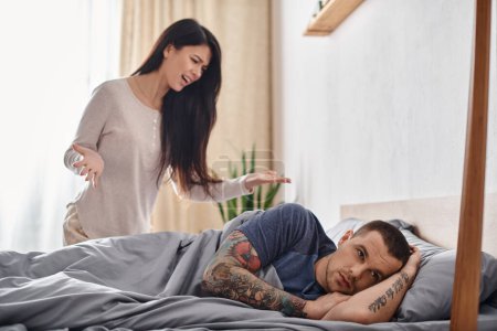Téléchargez les photos : Irrité asiatique femme querelle à offensé tatoué mari couché sur lit à la maison, divorce concept - en image libre de droit