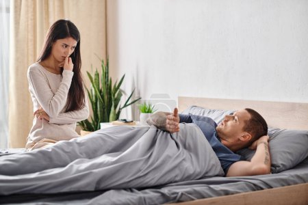Téléchargez les photos : Offensé asiatique femme regardant jeune tatoué mari couché sur lit à la maison, divorce concept - en image libre de droit