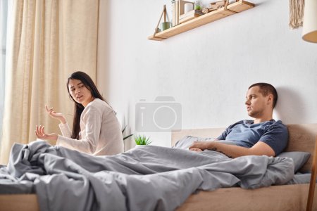 Photo for Offended asian woman talking to young tattooed husband lying on bed at home, divorce concept - Royalty Free Image