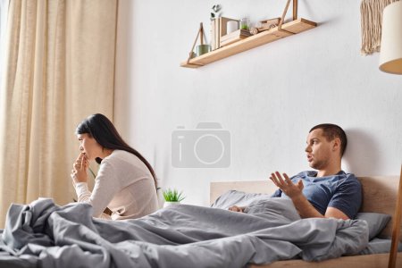 Photo for Young  man talking to offended asian woman while lying down in bedroom, relationship trouble - Royalty Free Image