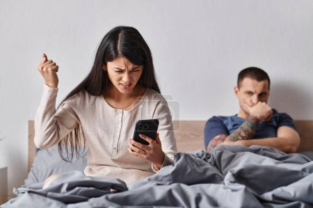 angry asian woman looking at smartphone near husband lying on bed at home, family conflict