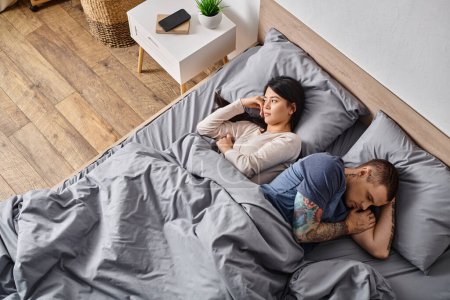 alto ángulo vista de asiático esposa y tatuado hombre acostado en la cama, relación dificultades concepto