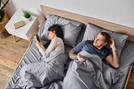 young asian woman looking in smartphone of husband lying on bed at home, family conflict concept