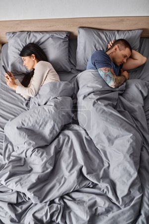 young tattooed man near asian wife looking at his smartphone in bedroom, misunderstanding, top view