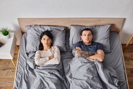 Ansicht von oben: junges frustriertes multikulturelles Paar legt sich zu Hause mit verschränkten Armen auf das Bett