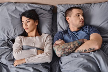 Draufsicht eines depressiven gemischtrassigen Paares, das sich mit verschränkten Armen auf das Bett legt und wegsieht