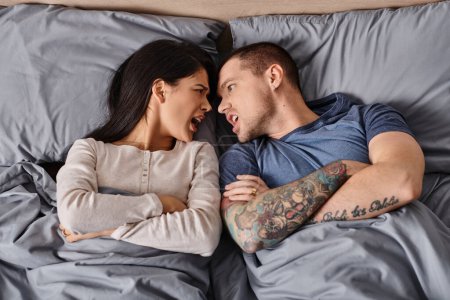 Téléchargez les photos : Vue de dessus du couple interracial en colère couché avec les bras croisés et se quereller sur le lit à la maison - en image libre de droit