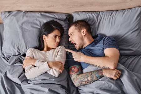 Téléchargez les photos : Vue de dessus du couple interracial irrité couché sur le lit criant et se querellant les uns aux autres - en image libre de droit