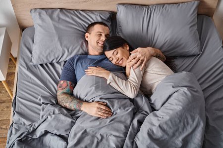 Téléchargez les photos : Vue de dessus de jeune couple multiculturel joyeux embrassant dans la chambre à coucher à la maison, bonheur familial - en image libre de droit