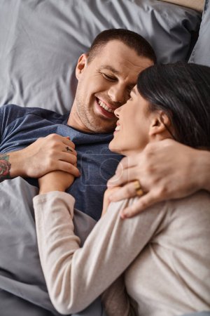 top view of young and joyous interracial couple embracing in bedroom at home, family happiness