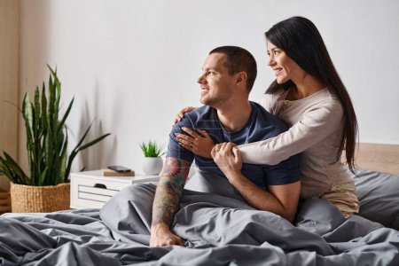 cheerful asian woman hugging young tattooed man looking away in bedroom at home, joy and happiness