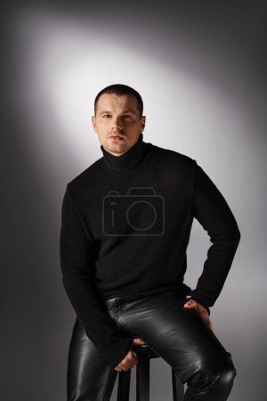 joven hombre de moda en cuello alto negro sentado y mirando a la cámara en gris con iluminación