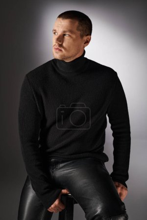 pensive trendy man in black turtleneck sitting and looking away on grey backdrop with lighting