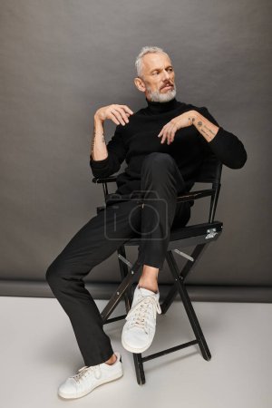 atractivo modelo masculino maduro en cuello alto de moda sentado en silla plegable y mirando hacia otro lado