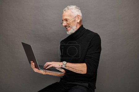 cheerful handsome bearded mature man with glasses sitting on chair with laptop and smiling happily