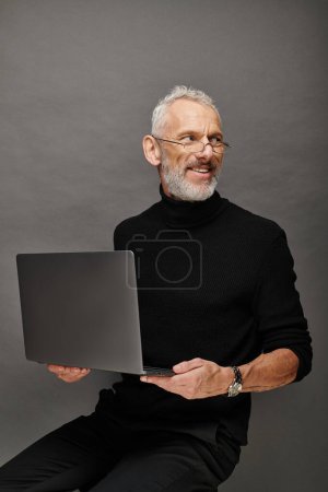 joyous attractive bearded mature man with glasses sitting on chair with laptop and smiling happily