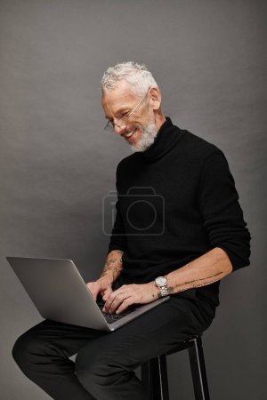 joyous attractive bearded mature man with glasses sitting on chair with laptop and smiling happily