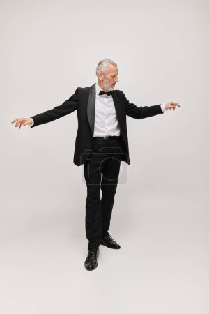 gut aussehendes reifes männliches Modell mit Fliege und Bart im eleganten Smoking, der auf grauem Hintergrund tanzt