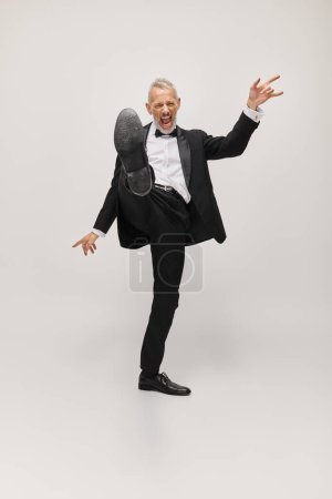 joyful good looking mature man in elegant suit with bow tie dancing happily on gray backdrop