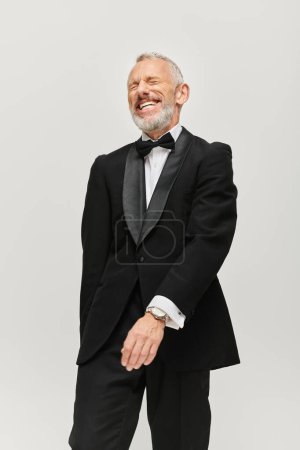 joyful good looking mature man with bow tie and gray beard in chic tuxedo smiling happily